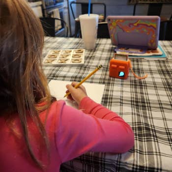 A child in a pink top writes on paper at a checkered table with educational tools and a tablet nearby