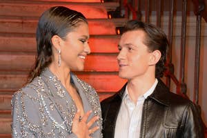 Two individuals stand on stairs; one wears a bejeweled blazer, the other a leather jacket. They gaze at each other, smiling at an event