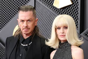 Two people on a red carpet; one wears a black suit, the other a sleeveless, patterned gown holding a clutch