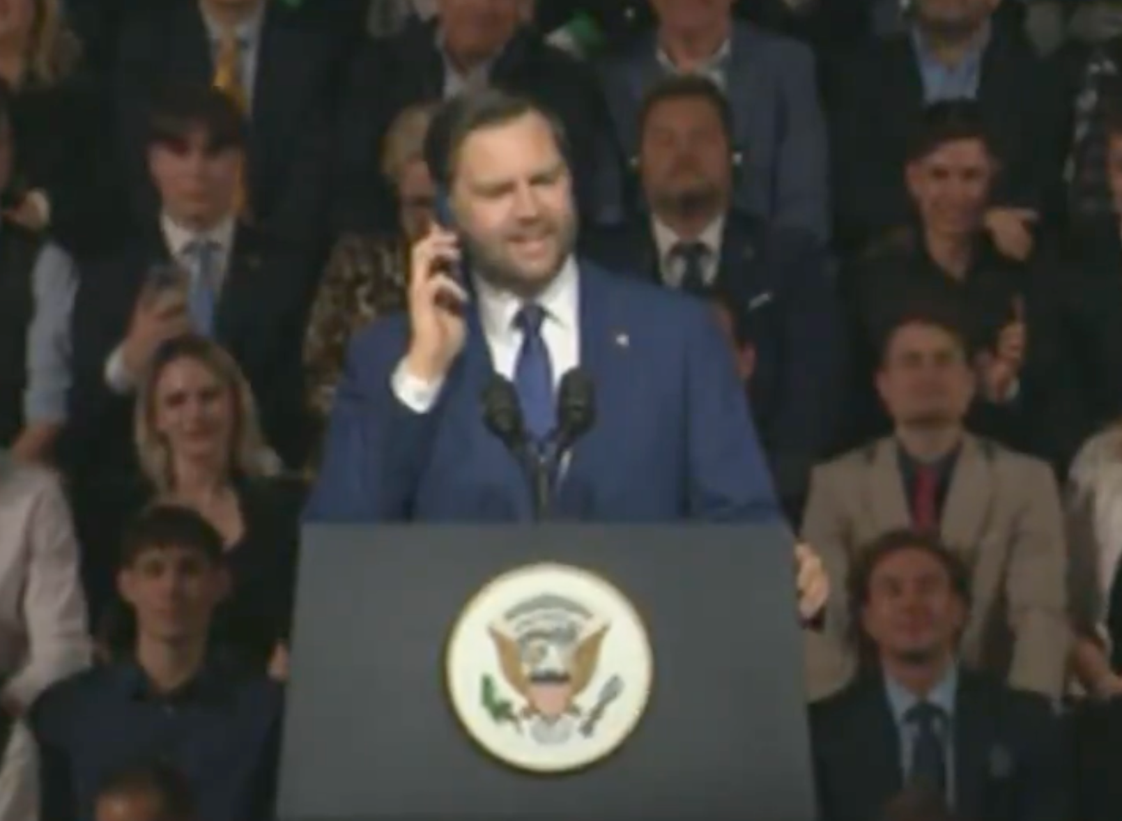 Person in a suit speaks at a podium with a presidential emblem, addressing an audience