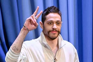 Person in casual tracksuit and boots makes a peace sign on a stage with blue curtains in the background