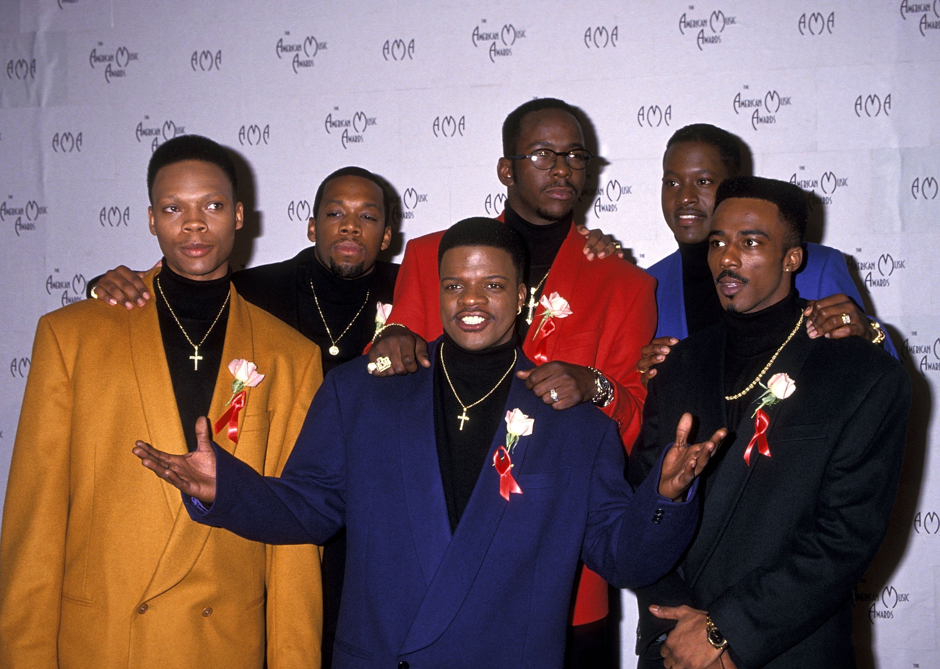 Group of men in stylish blazers with red ribbon pins at a formal event, posing together and smiling for the camera