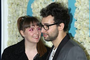 Two people posing closely at an event. One person wears a black jacket with sparkly makeup, and the other wears glasses and a gray suit jacket