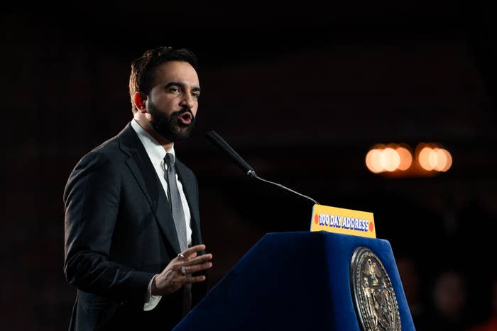 Person in a suit delivers a speech at a podium labeled "NYC 2023 Address," appearing focused and engaged with the audience