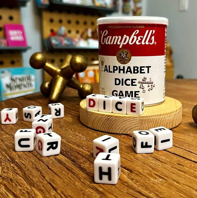 Campbell's Alphabet Dice Game set on a wooden table, surrounded by jacks and letters. Background features shelves with various boxed games