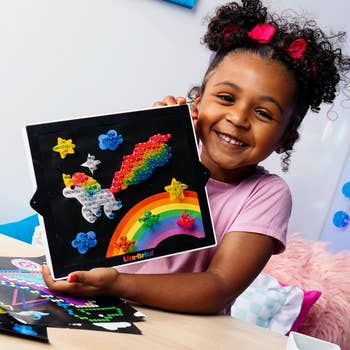 Child smiling and displaying a Lite-Brite unicorn and rainbow design on a black board, with colorful pegs