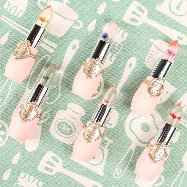 Six lipsticks with transparent bullet tips featuring embedded flowers and silver decorative accents on a patterned background