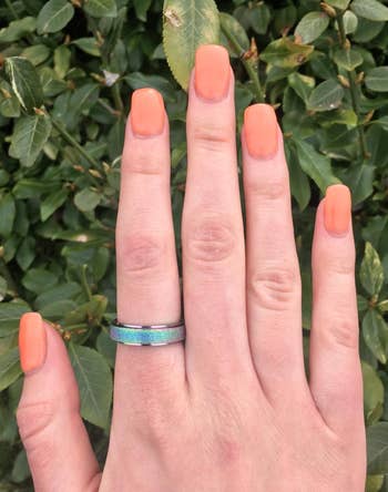 A hand with orange matte nails and a shiny blue-green ring is in front of green leaves