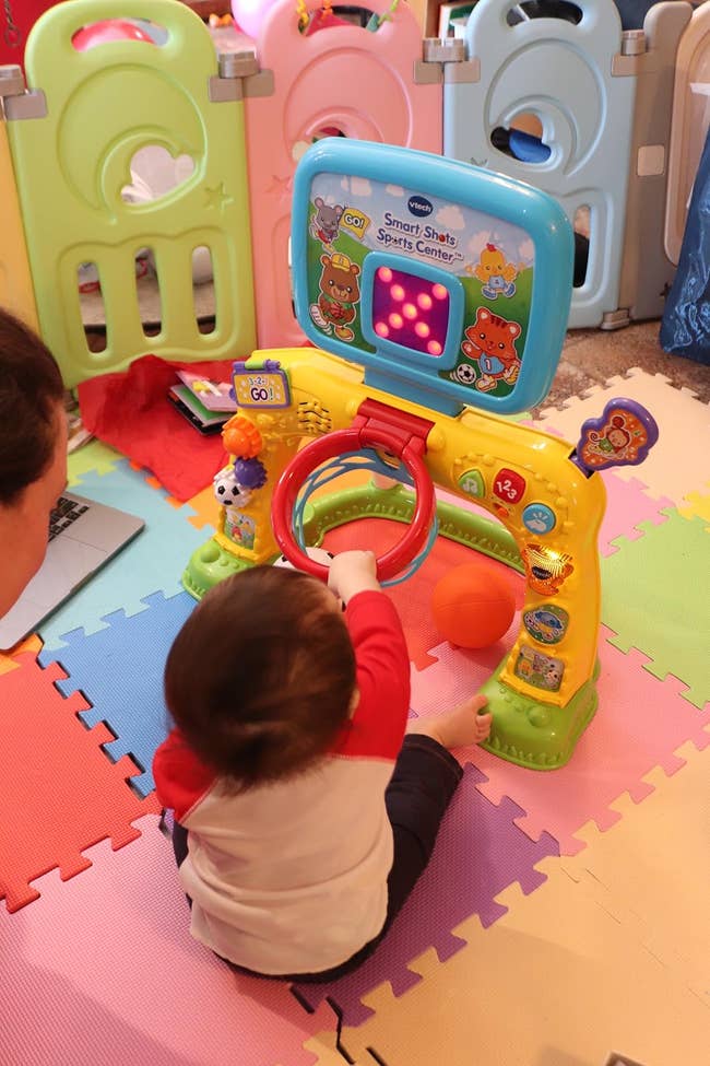 A child plays with an interactive toy featuring buttons, lights, and sports activities, seated on a colorful play mat