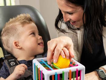 Child in a harnessed seat smiles at a woman holding a textured ball inside a sensory toy