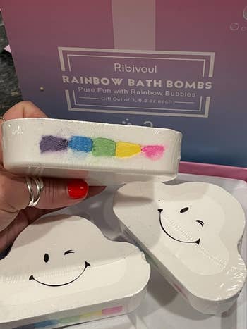 Person holding a cloud-shaped bath bomb with a colorful stripe. Background shows packaging for