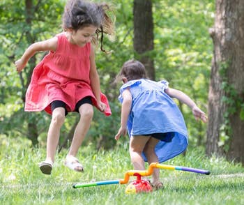 Two young children play in grass, jumping over a toy. One wears a pink dress, the other a blue dress. Trees are visible in the background