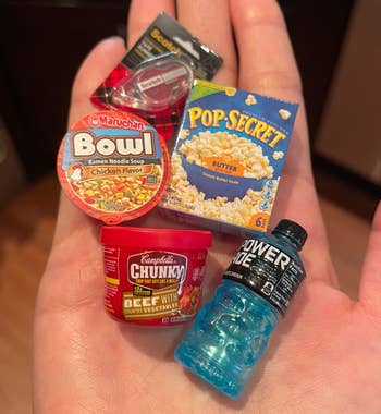 Hand holding five miniatures: Maruchan ramen cup, Pop-Secret popcorn box, Campbell's Chunky Beef with Vegetables soup, and a blue Powerade bottle