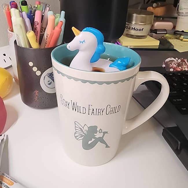 Unicorn-shaped tea infuser in a mug on a cluttered desk. The mug reads
