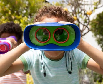 Child looking through toy binoculars outdoors, smiling with another child in the background. Ideal for exploring and learning toys
