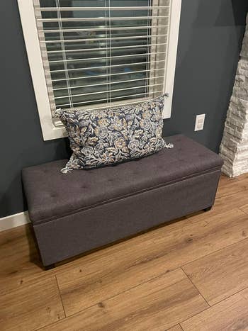 Tufted storage bench with a floral pillow, placed under a window in a modern living room setting