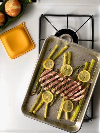 Baking tray with asparagus, lemon slices, and raw bacon arranged on a stovetop, next to onions and a square silicone mat
