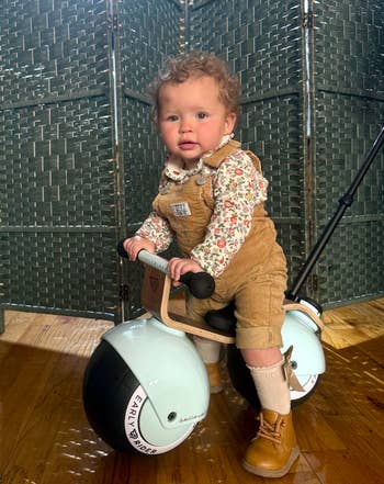 Toddler in floral shirt and corduroy overalls sits on a stylish ride-on toy indoors