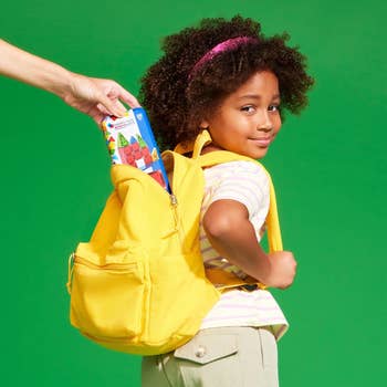 Child with yellow backpack getting a snack bar placed inside, smiling at the camera against a plain green background