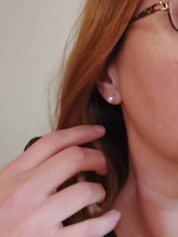 A close-up of a person's ear wearing a small pearl earring. A hand with light skin is near the ear, and the person is wearing glasses