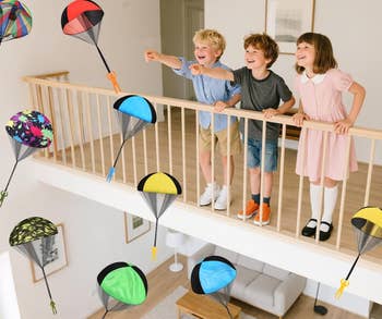 Three children on a balcony happily play with toy parachutes, which are floating down toward the floor below