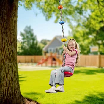Child swings on a rope swing attached to a tree in a sunny backyard with playground equipment in the background