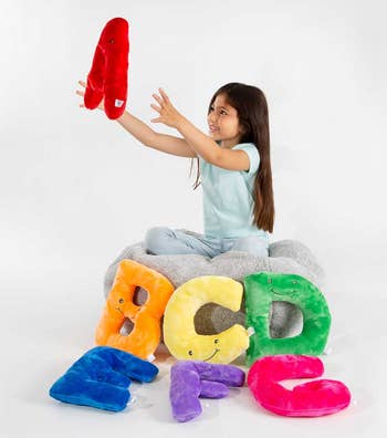 Child playing with letter-shaped plush toys, seated on a gray cushion. Plushies are styled in vibrant designs, ideal for a playful learning setting