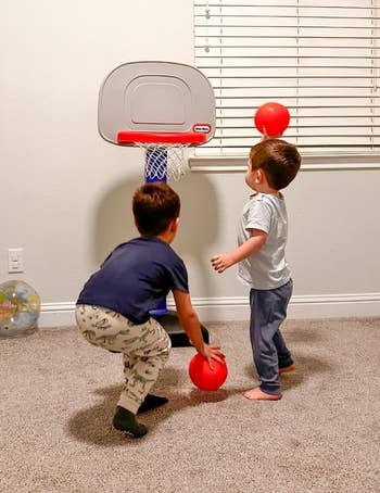 Two young children play with a toy basketball hoop indoors, each holding a small red ball, suggesting a fun, active playtime setup