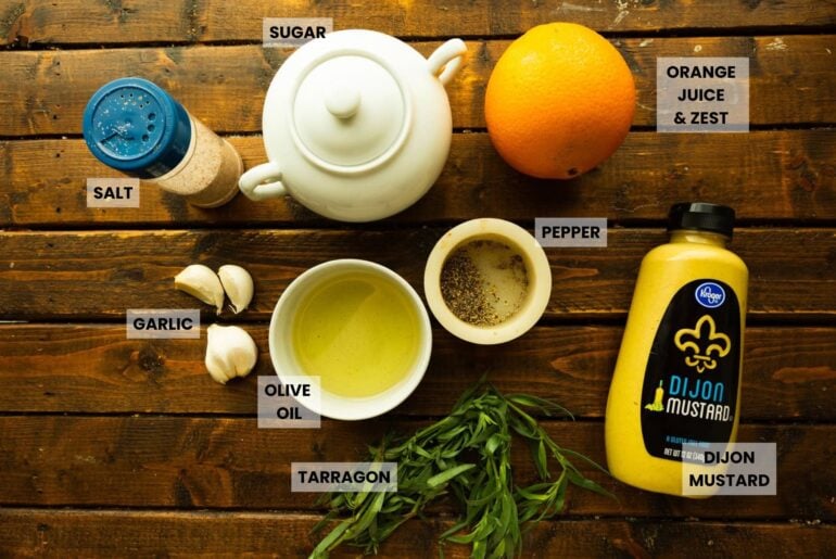 Flat lay of key ingredients: garlic cloves, olive oil, pepper, Dijon mustard, fresh tarragon, an orange, and a teapot, arranged on a rustic wood surface.