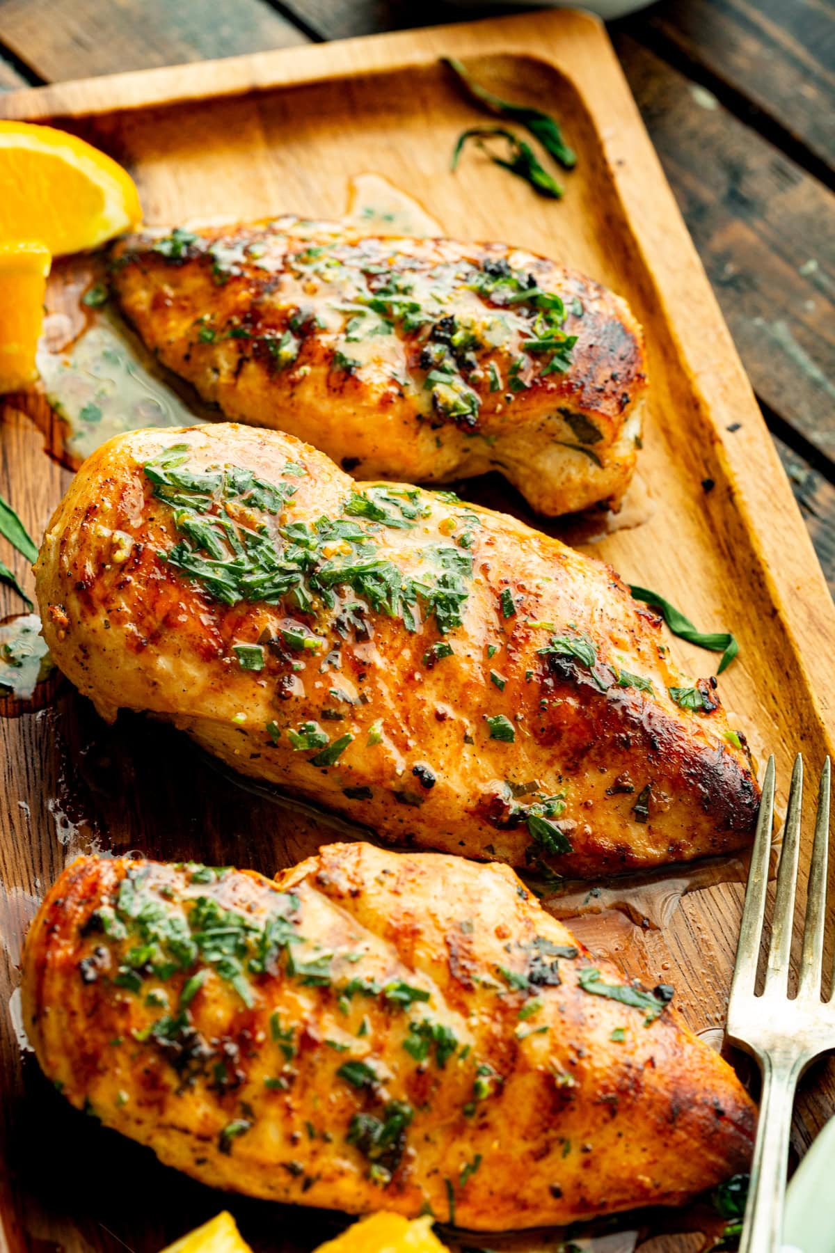 Grilled chicken breasts on a wooden tray with orange wedges, glistening with citrus marinade and fresh herbs.