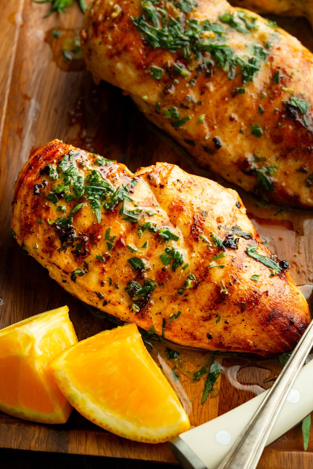 Multiple grilled chicken breasts on a wooden tray with orange wedges, glistening with citrus marinade and fresh herbs.