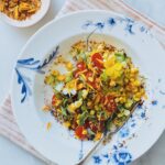 A colorful salad with corn, cherry tomatoes, cucumber, herbs, and edible flowers is served on a white plate with blue designs. A fork rests on the plate, and a small bowl of seasoning sits nearby on a striped cloth.