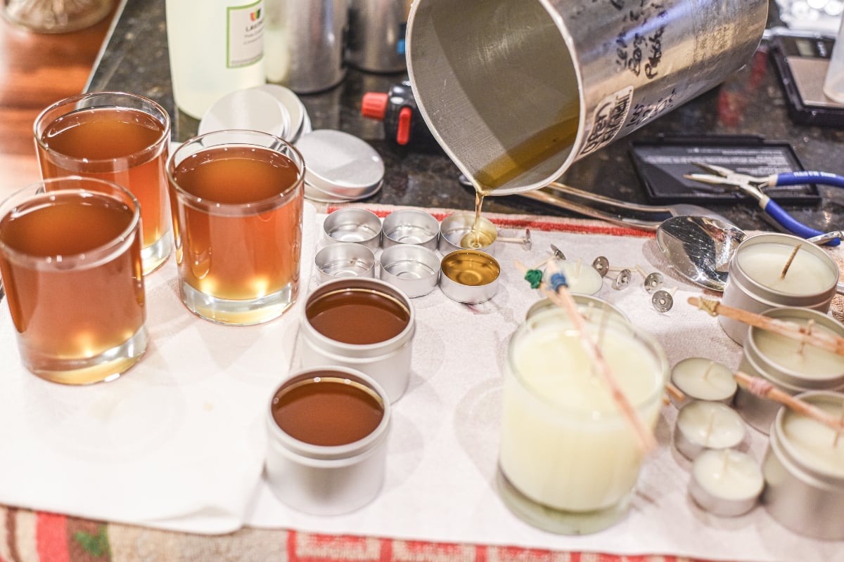 wax being poured from a metal container into tea lights with wicks surrounded by other candles in process