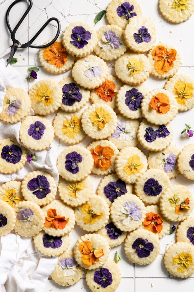 Cookies baked with edible flowers stacked on a white table.