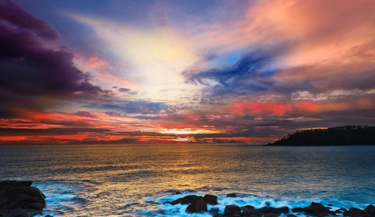 Colorful sunset over the ocean with waves crashing against rocks in the foreground and partly cloudy sky filled with vibrant hues of orange, pink, blue, and purple.
