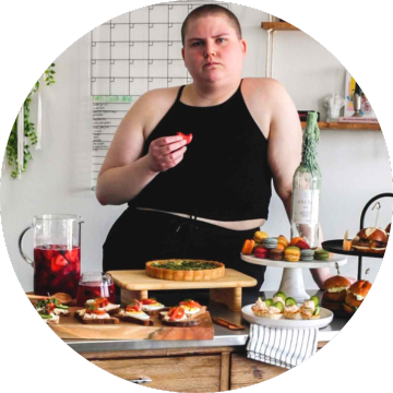 a person with a shaved head eating a macaron in front of a large table of party food