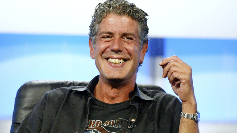 A smiling Anthony Bourdain sitting down with one arm raised near his face.