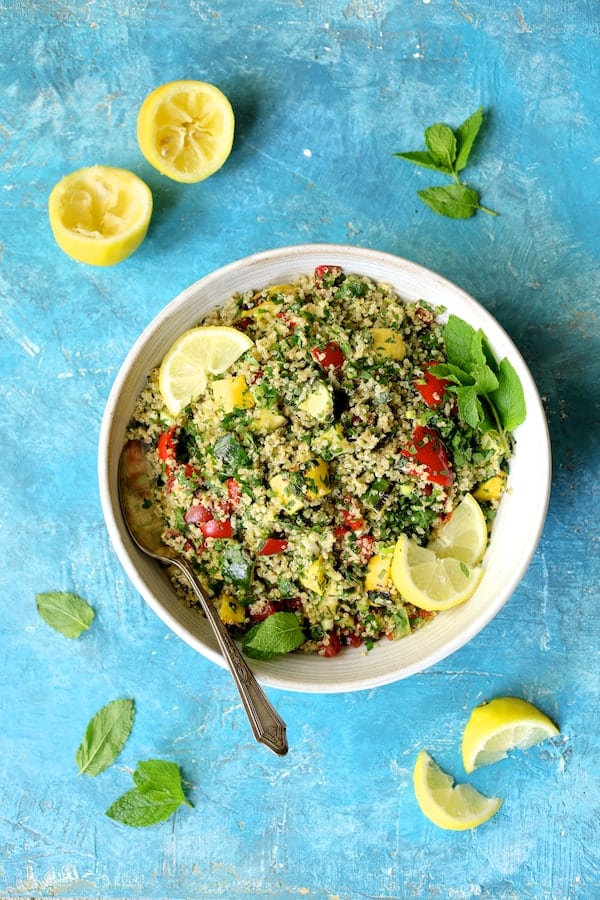 Photo of Quinoa Tabbouleh with Grilled Vegetables on blue background in white bowl garnished with mint leaves and lemon slices.