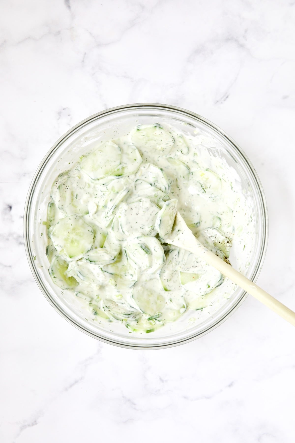 Cucumber salad being stirred in glass bowl with white spoon.