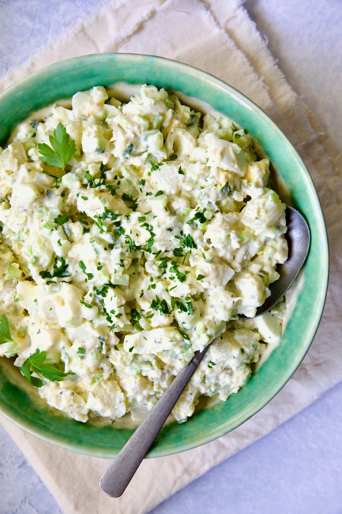 Creamy Potato Cucumber Salad in green and white serving bowl with serving spoon on frayed ivory towel.