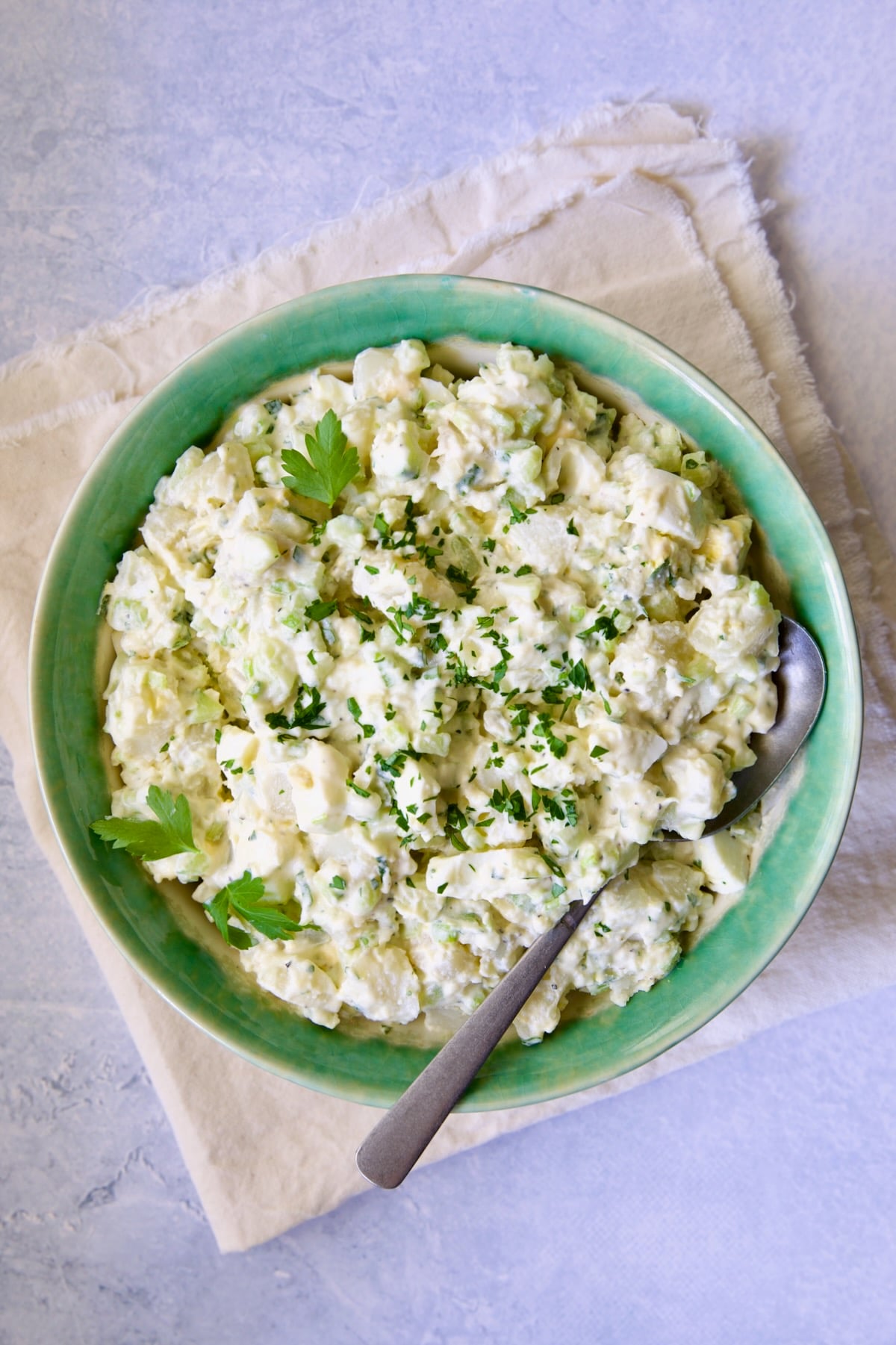 Creamy Potato Cucumber Salad in green and white serving bowl with serving spoon on frayed ivory towel.