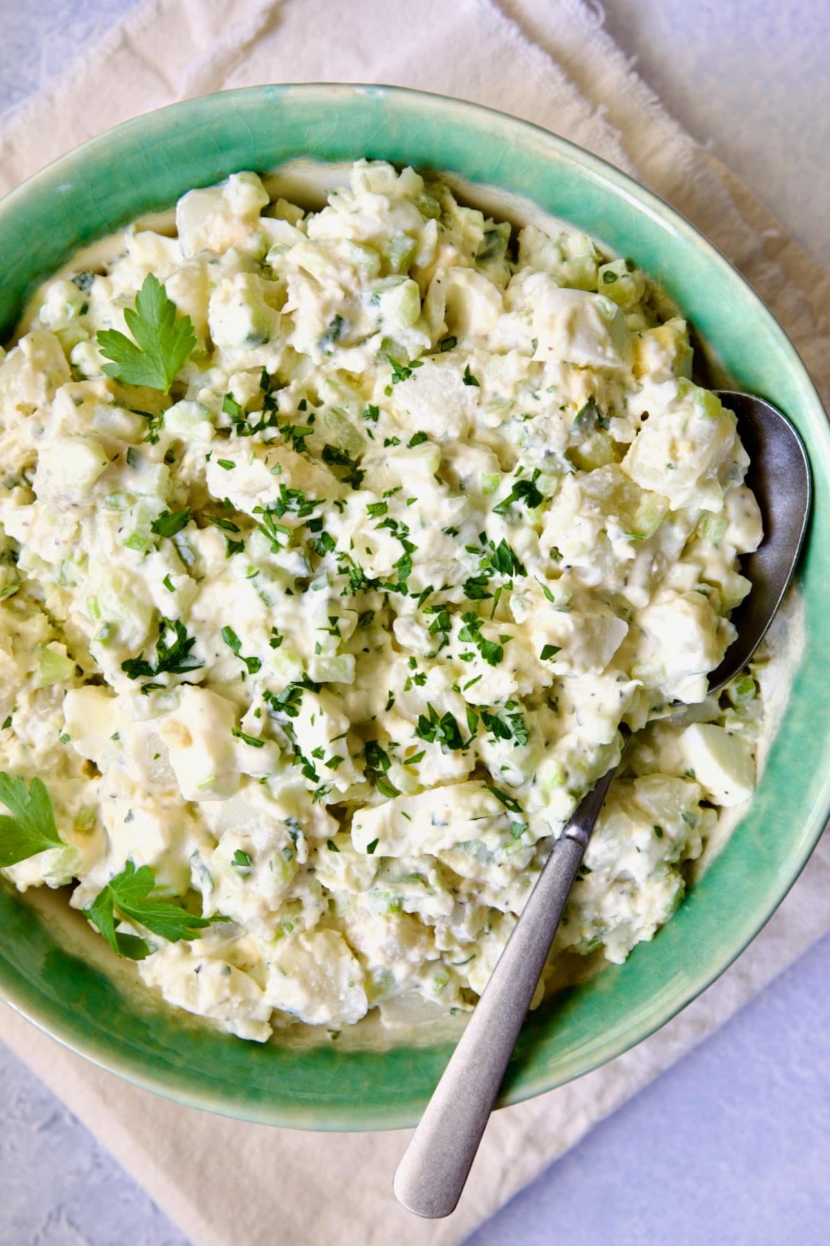 Creamy Potato Cucumber Salad in green and white serving bowl with serving spoon on frayed ivory towel.