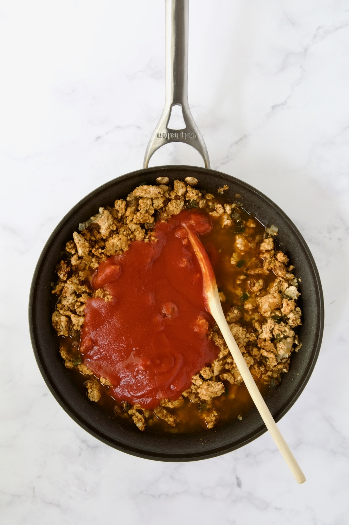 Tomato sauce added to ground chicken in nonstick skillet being stirred with wooden spoon.