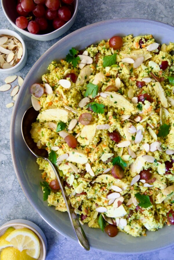 Chicken Couscous Salad in gray bowl with serving spoon with bowl of sliced almonds, whole grapes and lemon wedges.