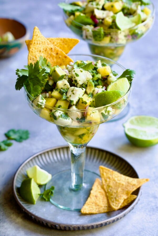 Halibut Ceviche with mango, avocado, lime wedges, cilantro sprigs and tortilla chips in margarita glass.