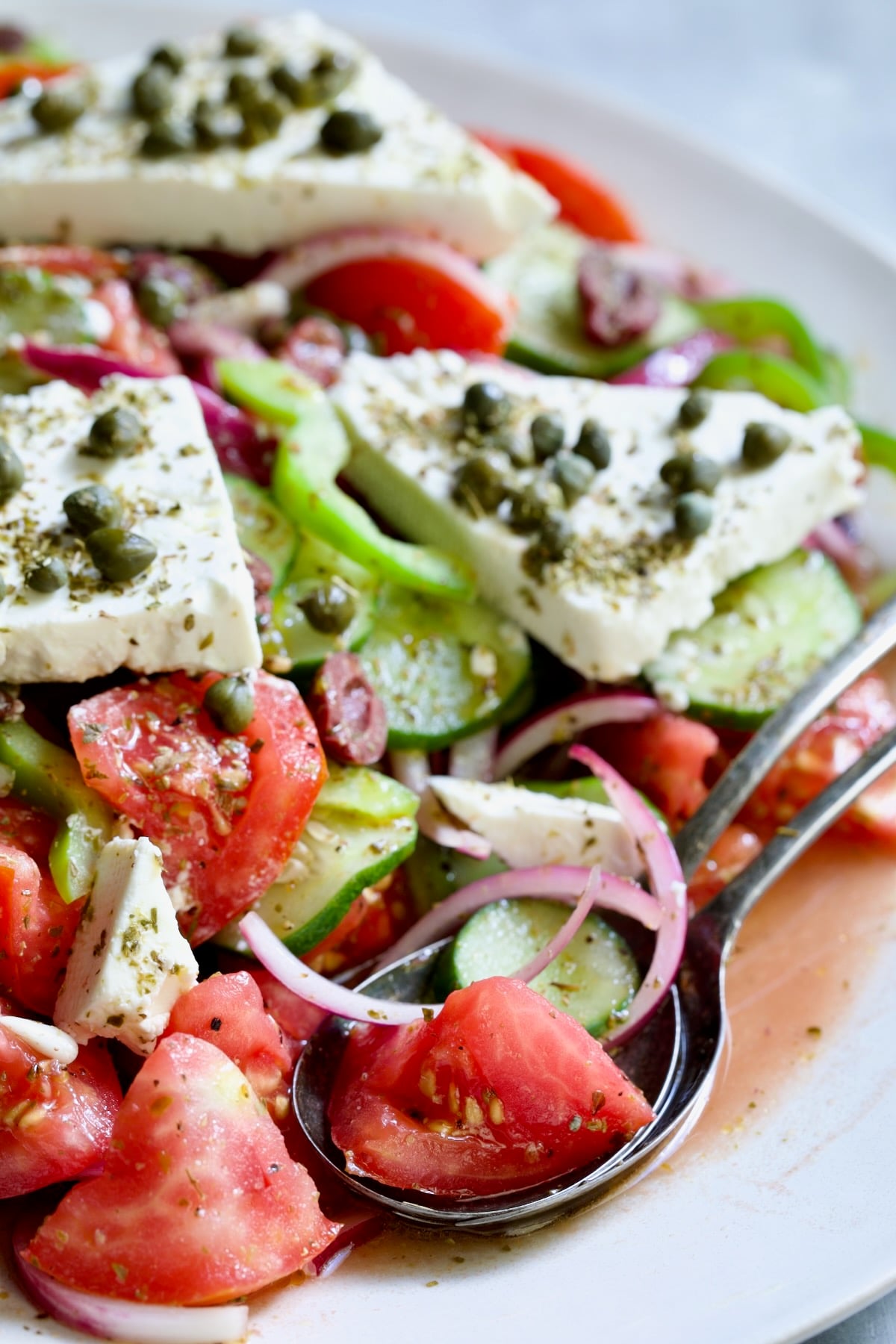 Classic Greek Salad with broken chunks of feta triangles on white oval platter with serving utensils.