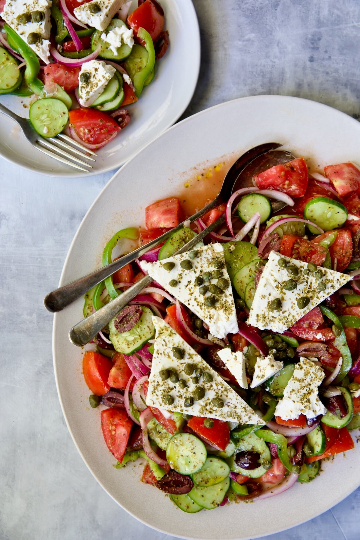 Classic Greek Salad with broken chunks of feta triangles on white oval platter with serving utensils with some served up on a small round plate.