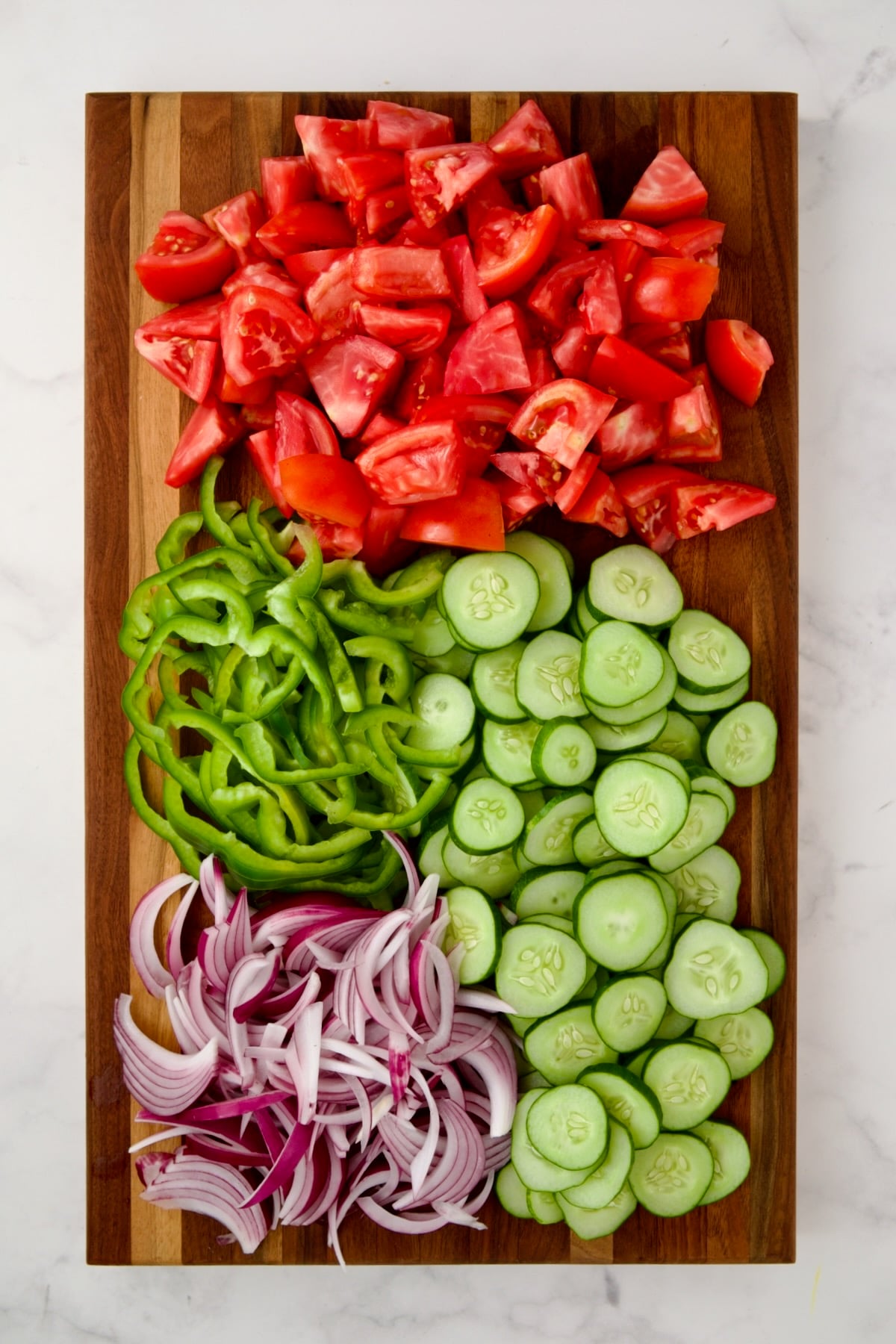 Chunks of tomatoes, sliced cucumbers, sliced green pepper and onion on wooden cutting board.