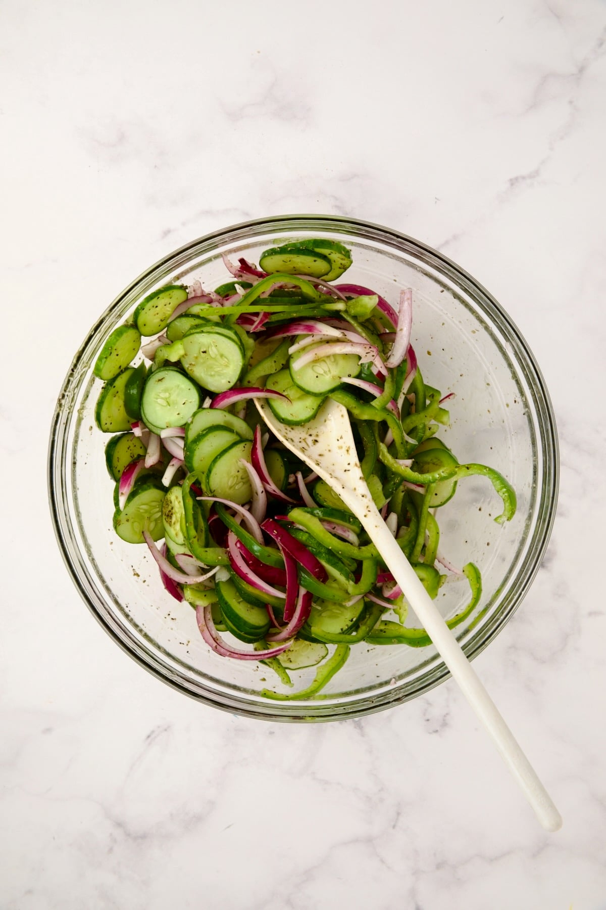 Sliced onion, green pepper and cucumbers in glass bowl being stirred with white mixing spoon.