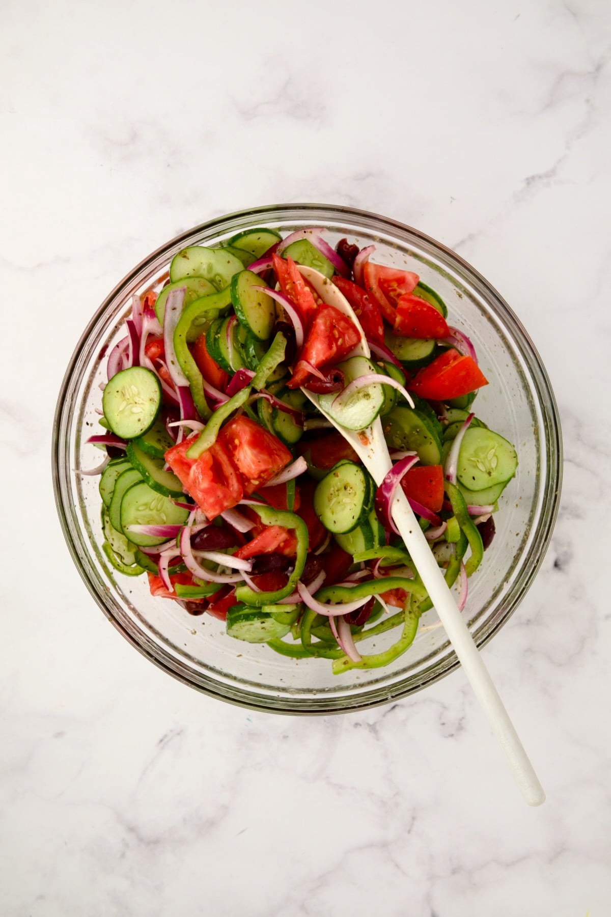 Tomatoes added to cucumbers, green pepper and onions in glass bowl being mixed with white mixing spoon.
